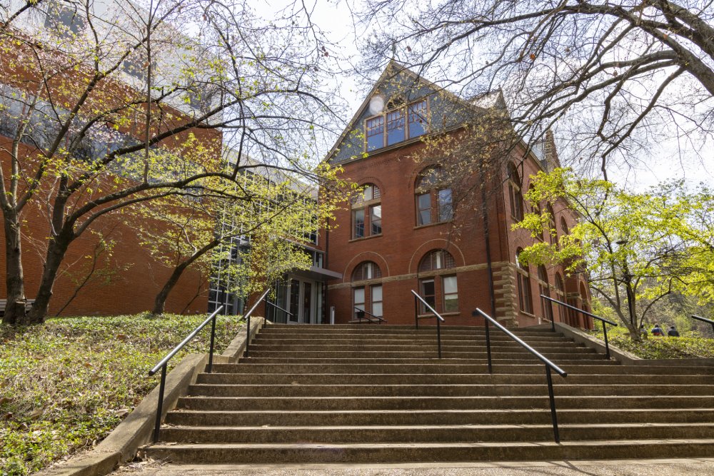 A red brick building with large arched windows and a steep triangular roof, framed by trees with budding green leaves. A wide set of stone stairs with black handrails leads up to the entrance, which features glass doors and large windows.